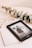 A Kindle device displaying a book titled 'Bridgerton: The Duke & I' rests on top of a closed book titled 'Five Min'. Beside the Kindle, there is a pair of eyeglasses with a gold frame and a sprig of eucalyptus leaves, all placed on a pale beige surface.