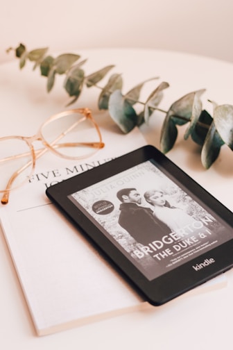 A Kindle device displaying a book titled 'Bridgerton: The Duke & I' rests on top of a closed book titled 'Five Min'. Beside the Kindle, there is a pair of eyeglasses with a gold frame and a sprig of eucalyptus leaves, all placed on a pale beige surface.