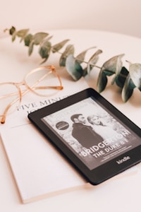 A Kindle device displaying a book titled 'Bridgerton: The Duke & I' rests on top of a closed book titled 'Five Min'. Beside the Kindle, there is a pair of eyeglasses with a gold frame and a sprig of eucalyptus leaves, all placed on a pale beige surface.