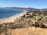 A scenic coastal landscape features a long stretch of sandy beach bordered by blue ocean waves. On one side, low vegetation and cacti cover a hillside, leading to a view of distant mountains and a town nestled along the coastline. Several houses and a small road with parked cars are visible in the middle distance.