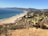 A scenic coastal landscape features a long stretch of sandy beach bordered by blue ocean waves. On one side, low vegetation and cacti cover a hillside, leading to a view of distant mountains and a town nestled along the coastline. Several houses and a small road with parked cars are visible in the middle distance.