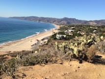 A scenic coastal landscape features a long stretch of sandy beach bordered by blue ocean waves. On one side, low vegetation and cacti cover a hillside, leading to a view of distant mountains and a town nestled along the coastline. Several houses and a small road with parked cars are visible in the middle distance.
