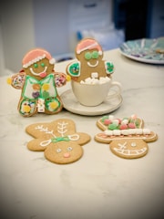 Three gingerbread cookies are decorated with icing and toppings. One is wearing a Santa hat and is sitting in a cup filled with marshmallows. Another gingerbread man stands next to the cup with colorful decorations. Two other cookies lie flat on the table, shaped like a reindeer and a face with a big grin, both featuring colorful icing details.