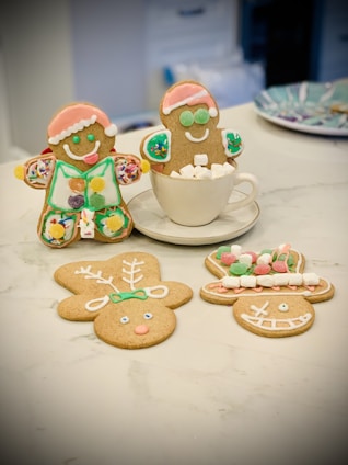 Three gingerbread cookies are decorated with icing and toppings. One is wearing a Santa hat and is sitting in a cup filled with marshmallows. Another gingerbread man stands next to the cup with colorful decorations. Two other cookies lie flat on the table, shaped like a reindeer and a face with a big grin, both featuring colorful icing details.