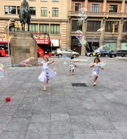 Three children are joyfully chasing large soap bubbles in an open urban plaza. Two girls run forward with outstretched arms, while a third child in the background also participates in the playful activity. Behind them, a busy street with a red double-decker bus and several parked cars can be seen. A statue of a person on horseback is situated on a pedestal nearby, with tall buildings forming the backdrop.