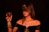 A woman with dramatic black makeup around her eyes and lips holds a decorative goblet filled with a red liquid. She is wearing an off-the-shoulder black lace dress, and the background is dimly lit, creating a moody and mysterious atmosphere.