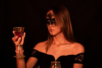 A woman with dramatic black makeup around her eyes and lips holds a decorative goblet filled with a red liquid. She is wearing an off-the-shoulder black lace dress, and the background is dimly lit, creating a moody and mysterious atmosphere.