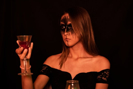A woman with dramatic black makeup around her eyes and lips holds a decorative goblet filled with a red liquid. She is wearing an off-the-shoulder black lace dress, and the background is dimly lit, creating a moody and mysterious atmosphere.