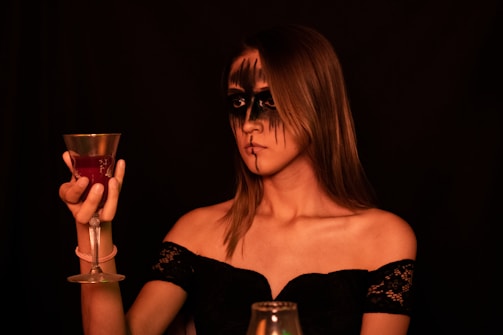 A woman with dramatic black makeup around her eyes and lips holds a decorative goblet filled with a red liquid. She is wearing an off-the-shoulder black lace dress, and the background is dimly lit, creating a moody and mysterious atmosphere.