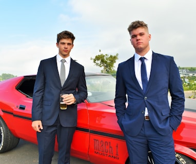 two men in suits standing next to a red car