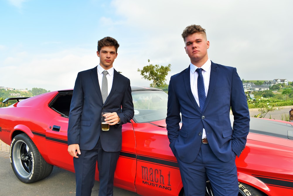 two men in suits standing next to a red car