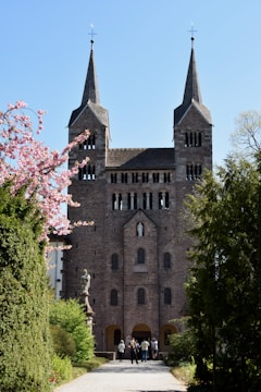 A grand stone church with two tall spires stands in a serene setting surrounded by lush greenery and a flowering tree. Several people are walking towards the entrance, and a statue of a religious figure is visible to the left of the path.