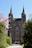 A grand stone church with two tall spires stands in a serene setting surrounded by lush greenery and a flowering tree. Several people are walking towards the entrance, and a statue of a religious figure is visible to the left of the path.