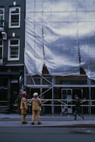 Two people dressed in protective gear stand in front of a construction site covered with white tarpaulin. The building is undergoing maintenance or renovation, evidenced by the scaffold and protective covering. Nearby, pedestrians walk by, casually observing the scene.