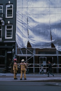 Two people dressed in protective gear stand in front of a construction site covered with white tarpaulin. The building is undergoing maintenance or renovation, evidenced by the scaffold and protective covering. Nearby, pedestrians walk by, casually observing the scene.