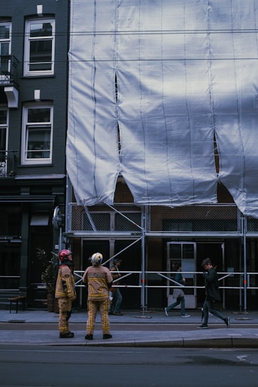 Two people dressed in protective gear stand in front of a construction site covered with white tarpaulin. The building is undergoing maintenance or renovation, evidenced by the scaffold and protective covering. Nearby, pedestrians walk by, casually observing the scene.
