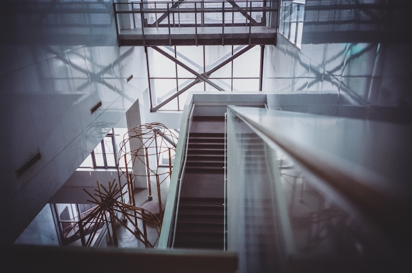 A modern interior with a geometric architectural design featuring a staircase descending through an open atrium. The space is framed by large windows allowing natural light to cast intricate shadows on the walls. An abstract metal sculpture is visible in the lower section, adding an artistic element to the sleek and minimalist design.