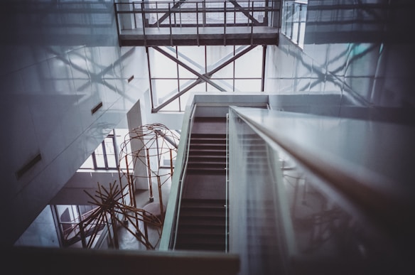 A modern interior with a geometric architectural design featuring a staircase descending through an open atrium. The space is framed by large windows allowing natural light to cast intricate shadows on the walls. An abstract metal sculpture is visible in the lower section, adding an artistic element to the sleek and minimalist design.