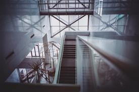 A modern interior with a geometric architectural design featuring a staircase descending through an open atrium. The space is framed by large windows allowing natural light to cast intricate shadows on the walls. An abstract metal sculpture is visible in the lower section, adding an artistic element to the sleek and minimalist design.