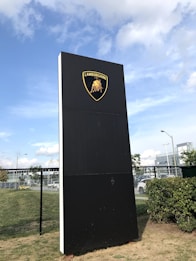 A tall black sign prominently features the Lamborghini logo with a bull emblem set against a clear blue sky. The sign is positioned on a grassy area with a chain-link fence in the background, along with some green bushes. In the distance, vehicles and an Acura building are visible.