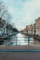 A scenic canal lined with trees runs through a city with traditional European-style buildings on both sides. A red bicycle is parked on the brick-paved bridge overlooking the canal, adding a pop of color amidst the muted tones of the urban landscape.