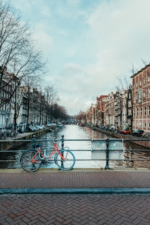 A scenic canal lined with trees runs through a city with traditional European-style buildings on both sides. A red bicycle is parked on the brick-paved bridge overlooking the canal, adding a pop of color amidst the muted tones of the urban landscape.