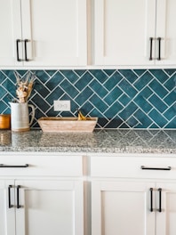 A kitchen countertop with white cabinets and black handles is set against a backdrop of teal herringbone tiles. The countertop holds a ceramic jug with dried flowers, a wooden tray with bananas, and another small container.