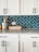 A kitchen countertop with white cabinets and black handles is set against a backdrop of teal herringbone tiles. The countertop holds a ceramic jug with dried flowers, a wooden tray with bananas, and another small container.