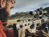 A person is carefully aiming a precision rifle equipped with various optics and mounted on a bipod. The setting appears to be a natural outdoor environment with blurred mountains in the background. The person is holding the rifle with focus, indicating preparation for a shot or practice.