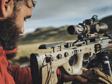 A person is carefully aiming a precision rifle equipped with various optics and mounted on a bipod. The setting appears to be a natural outdoor environment with blurred mountains in the background. The person is holding the rifle with focus, indicating preparation for a shot or practice.