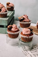 Cupcakes topped with pink frosting and garnished with nuts and chocolate, displayed on glass surfaces. They are arranged creatively with some cupcakes elevated on clear glass cups and others placed on a wooden crate.