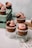 Cupcakes topped with pink frosting and garnished with nuts and chocolate, displayed on glass surfaces. They are arranged creatively with some cupcakes elevated on clear glass cups and others placed on a wooden crate.