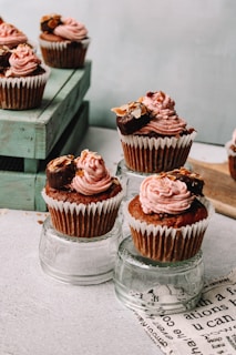 Cupcakes topped with pink frosting and garnished with nuts and chocolate, displayed on glass surfaces. They are arranged creatively with some cupcakes elevated on clear glass cups and others placed on a wooden crate.