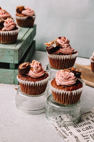 Cupcakes topped with pink frosting and garnished with nuts and chocolate, displayed on glass surfaces. They are arranged creatively with some cupcakes elevated on clear glass cups and others placed on a wooden crate.