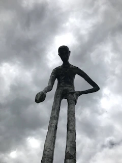A lone figure stands amid towering biomechanical ribs under a neon purple sky, with a skeletal giant's hand resting gently on their shoulder.