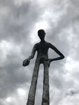 A lone figure stands amid towering biomechanical ribs under a neon purple sky, with a skeletal giant's hand resting gently on their shoulder.