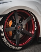 A close-up view of a customized car wheel featuring a carbon fiber rim with red accents. The tire has prominent white lettering and the brake caliper is in a contrasting orange shade.