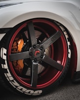A close-up view of a customized car wheel featuring a carbon fiber rim with red accents. The tire has prominent white lettering and the brake caliper is in a contrasting orange shade.