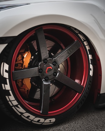 A close-up view of a customized car wheel featuring a carbon fiber rim with red accents. The tire has prominent white lettering and the brake caliper is in a contrasting orange shade.
