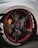 A close-up view of a customized car wheel featuring a carbon fiber rim with red accents. The tire has prominent white lettering and the brake caliper is in a contrasting orange shade.