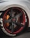 A close-up view of a customized car wheel featuring a carbon fiber rim with red accents. The tire has prominent white lettering and the brake caliper is in a contrasting orange shade.