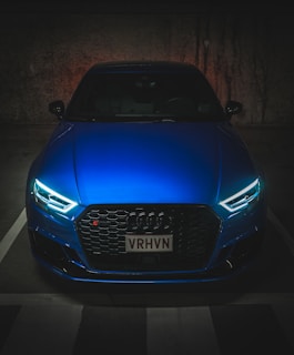 A sleek, modern car with illuminated headlights is parked in a dimly lit space. The car has a vivid blue color, and it features a distinctive grille with the manufacturer’s emblem prominently displayed.