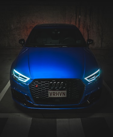 A sleek, modern car with illuminated headlights is parked in a dimly lit space. The car has a vivid blue color, and it features a distinctive grille with the manufacturer’s emblem prominently displayed.