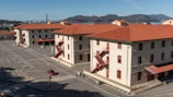 A cluster of three-story buildings with pale walls and red-tiled roofs is situated near a parking lot. The buildings feature red metal staircases on their exteriors. In the background, the iconic Golden Gate Bridge and hilly landscape are visible under a clear blue sky.