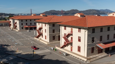 A cluster of three-story buildings with pale walls and red-tiled roofs is situated near a parking lot. The buildings feature red metal staircases on their exteriors. In the background, the iconic Golden Gate Bridge and hilly landscape are visible under a clear blue sky.