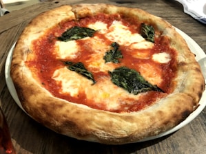 A classic Margherita pizza with a browned, puffy crust. The topping includes melted mozzarella cheese, vibrant red tomato sauce, and scattered fresh basil leaves. It is presented on a white plate set on a rustic wooden table, suggesting a casual dining setting.
