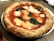 A classic Margherita pizza with a browned, puffy crust. The topping includes melted mozzarella cheese, vibrant red tomato sauce, and scattered fresh basil leaves. It is presented on a white plate set on a rustic wooden table, suggesting a casual dining setting.