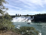 A majestic waterfall cascades into a wide river, creating mist and spray. In the foreground, lush green foliage frames the scene, while a boat filled with tourists approaches the falls. Skyscrapers and a blue sky are visible in the background.