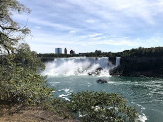 A majestic waterfall cascades into a wide river, creating mist and spray. In the foreground, lush green foliage frames the scene, while a boat filled with tourists approaches the falls. Skyscrapers and a blue sky are visible in the background.