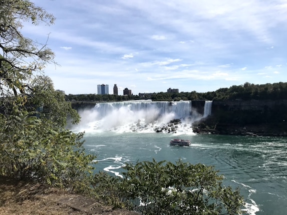 A majestic waterfall cascades into a wide river, creating mist and spray. In the foreground, lush green foliage frames the scene, while a boat filled with tourists approaches the falls. Skyscrapers and a blue sky are visible in the background.